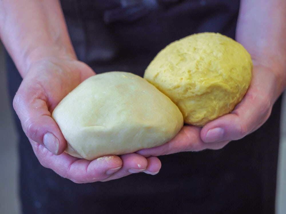 Hands holding two types of pasta dough balls, one is light and one is more yellow from the type of eggs.