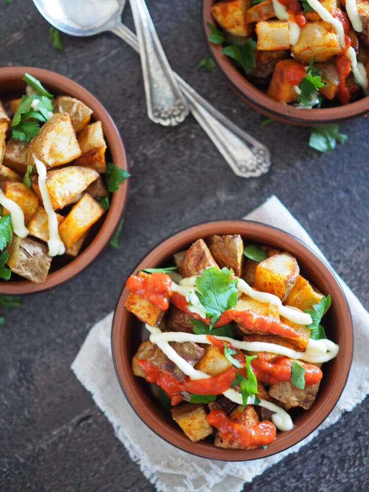 Patatas Bravas, a set of three terracotta dishes filled with oven baked potatoes, drizzled in salsa brava and aioli, and sprinkled with parsley.