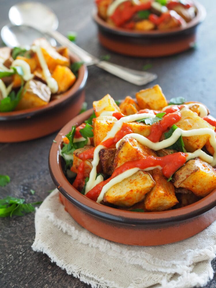 Patatas Bravas, a set of three terracotta dishes filled with oven baked potatoes, drizzled in salsa brava and aioli, and sprinkled with parsley.