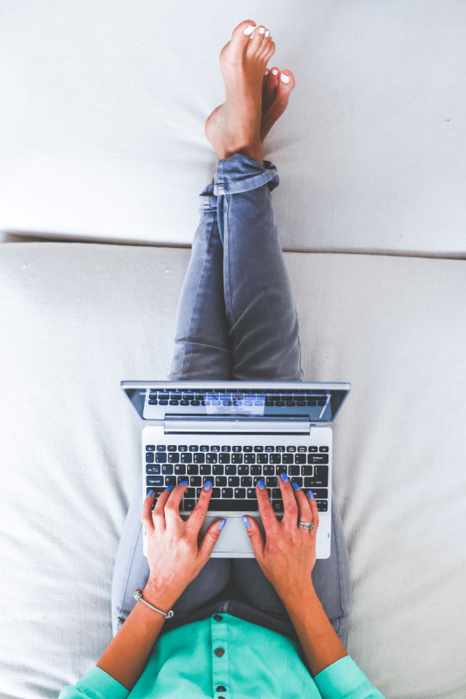 Overhead photo of girl in turquoise shirt resting on bed with legs crossed and typing on Macbook pro in lap planning future travel.