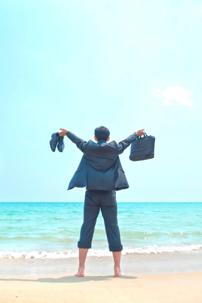 Man on beach facing ocean, wearing suit and holding up briefcase and dress shoes.