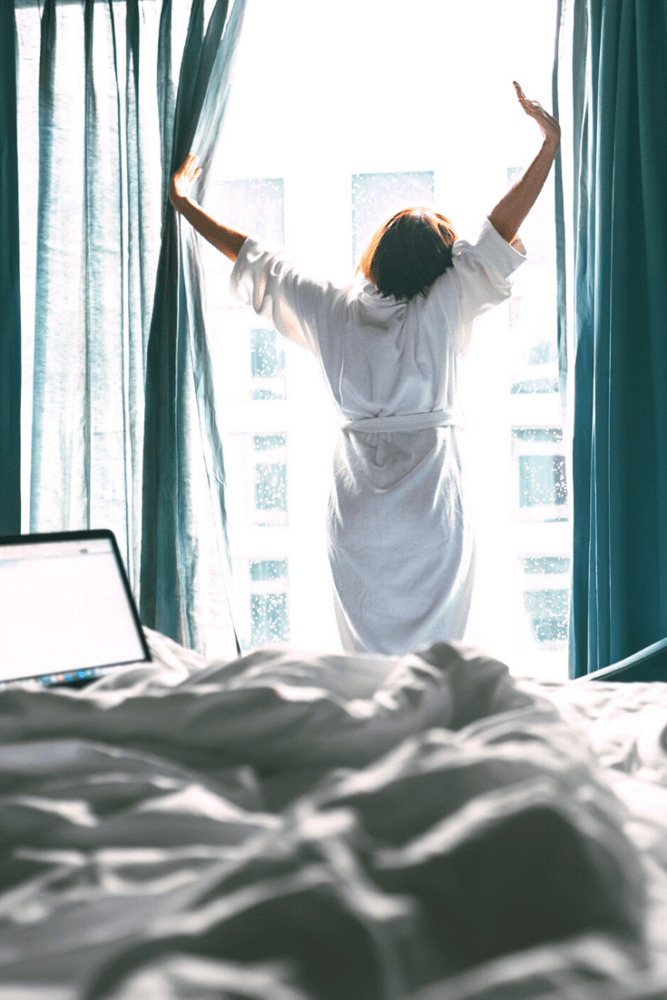 The Future Of Travel: Work Vacations. Woman in robe opening hotel curtains with laptop on unmade bed.