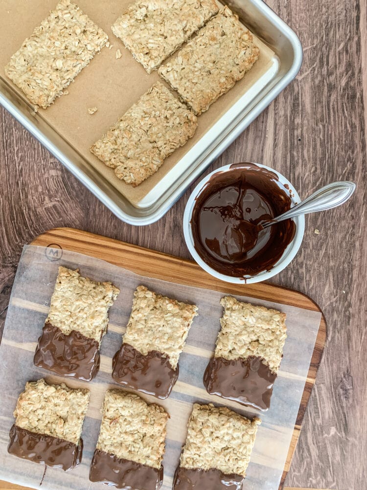 Baked oatcakes on a cookie sheet with a bowl of melted chocolate and chocolate dipped oatcakes cooling on wax paper.