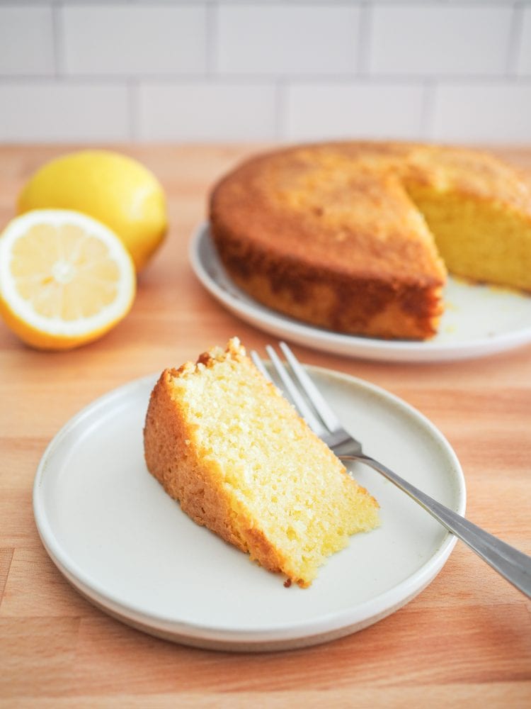 A slice of lemon olive oil cake on a plate with the whole cake in the background.