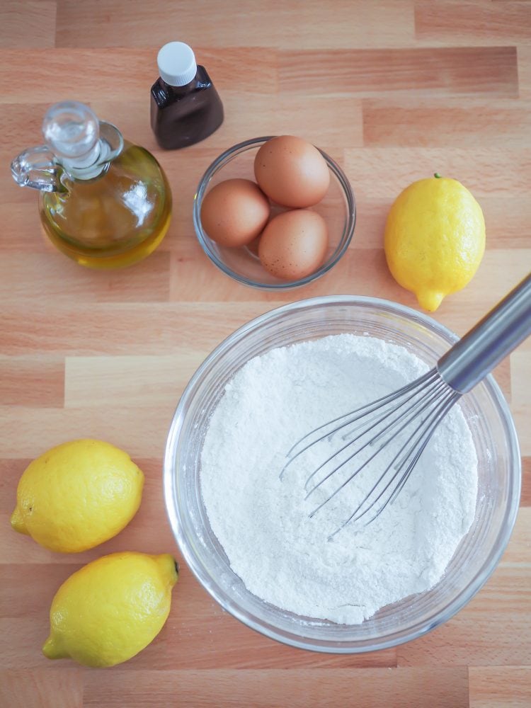 Overhead shot of mixing together the flour and baking powder in a bowl with eggs, oil, and lemons in the background.