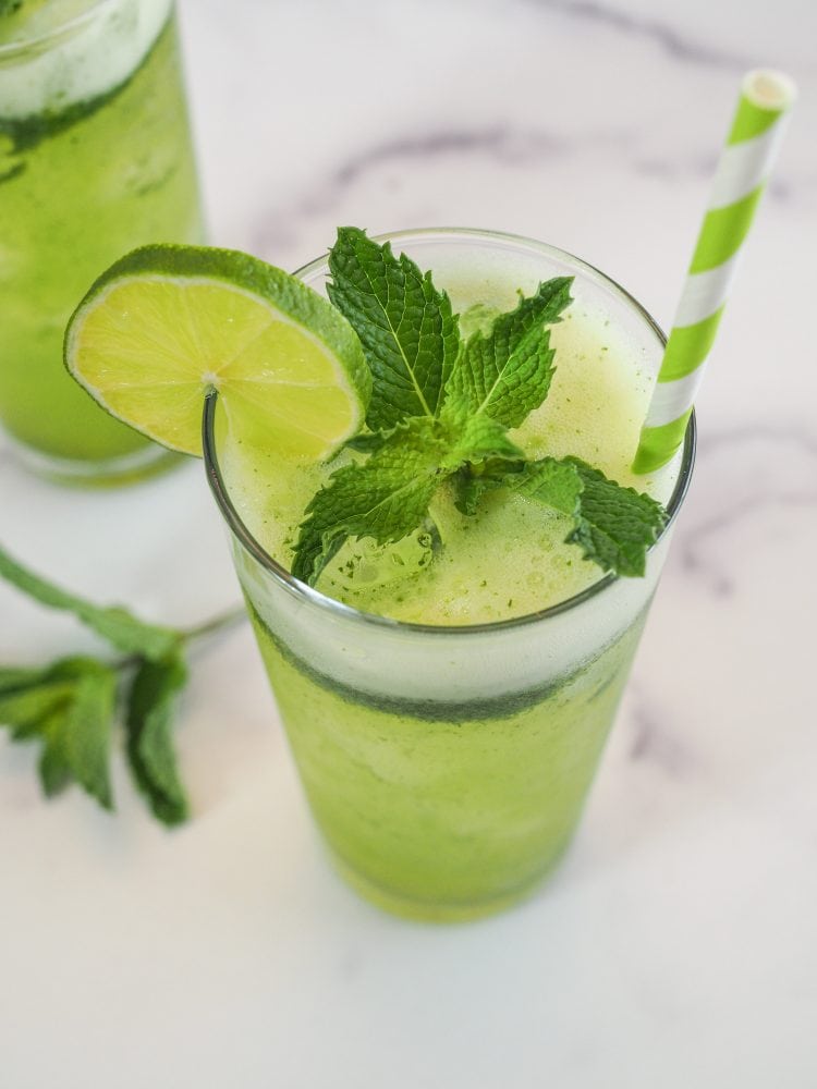 A glass of mint lemonade with slice of lime and sprig of mint as garnish, and a green and white paper straw.