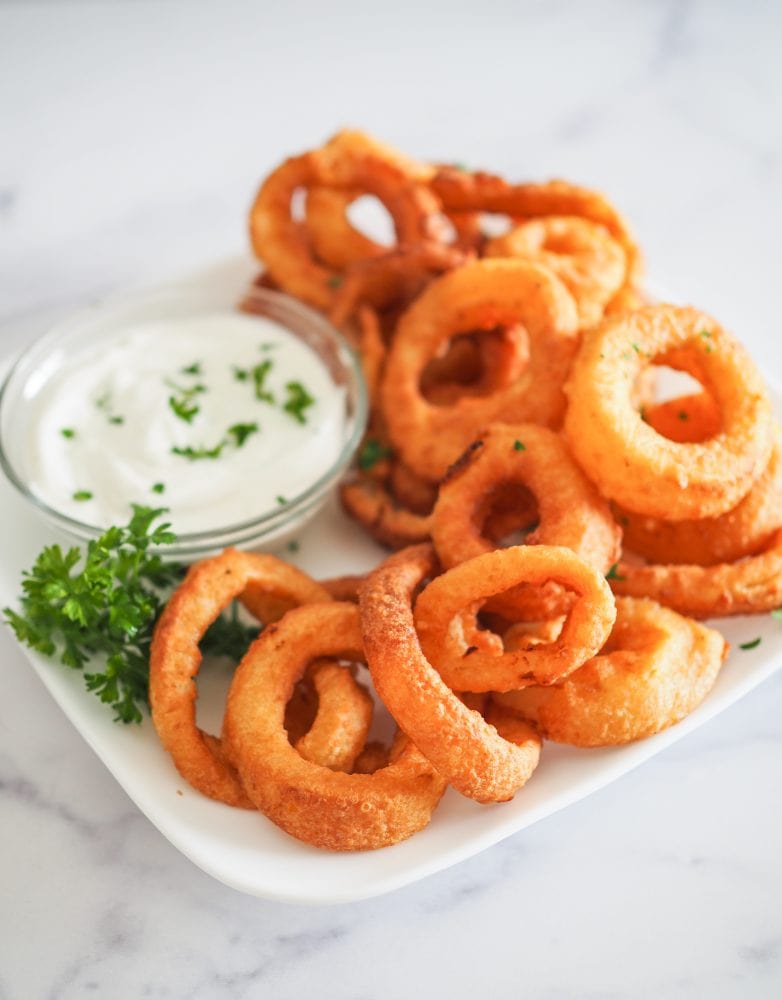 Side view of plate of air fried onion rings with a side of garlic dipping sauce and parsley.