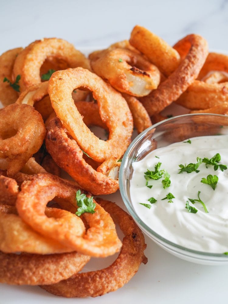 A close up of a plate of onion rings and a side of garlic dipping sauce with parsley.