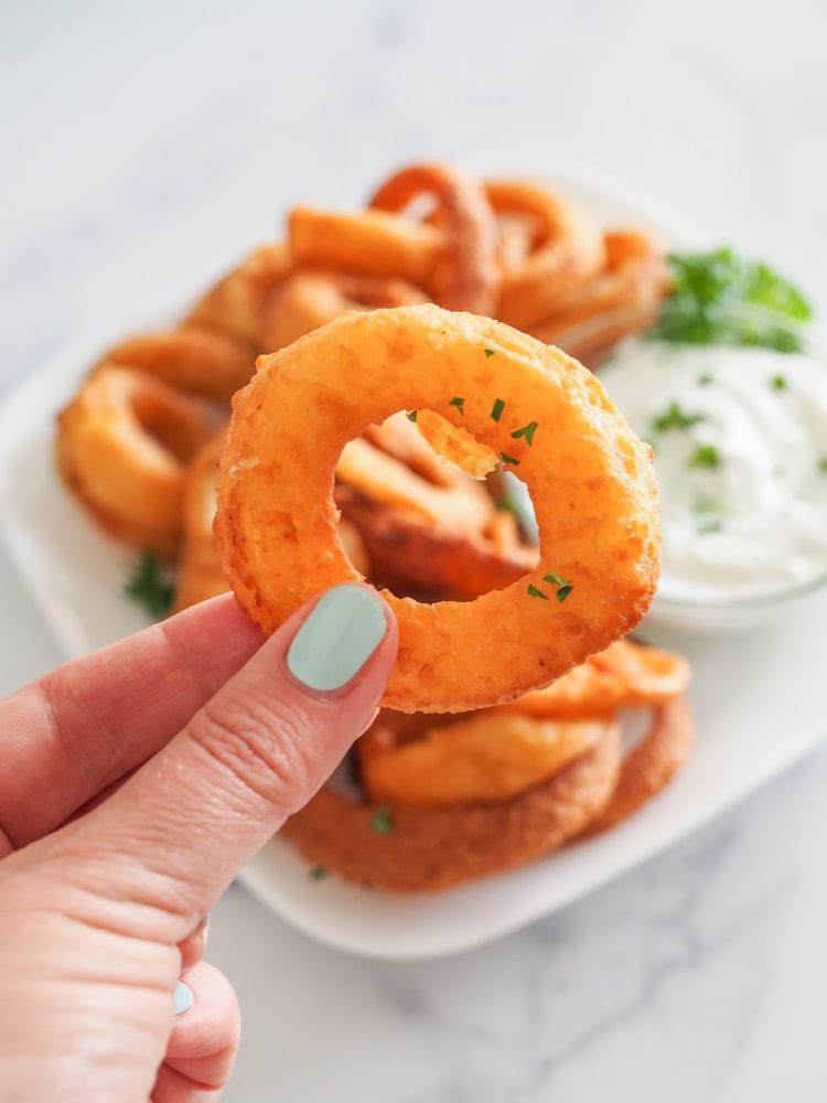 Fingers holding up a single round golden air fried onion ring with a plate of onion rings in the background.