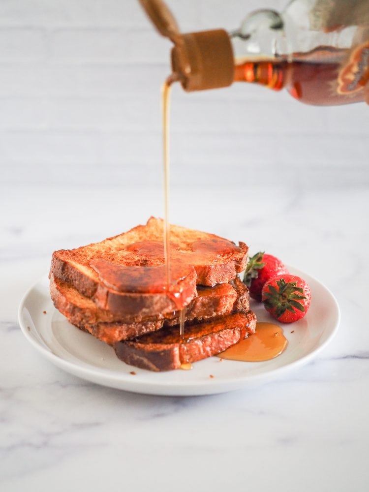 Pouring maple syrup onto four slices of french toast with strawberries on the side.