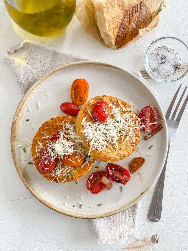 Overhead look at a plate with two round polenta cakes topped with Parmesan cheese and cherry tomatoes.