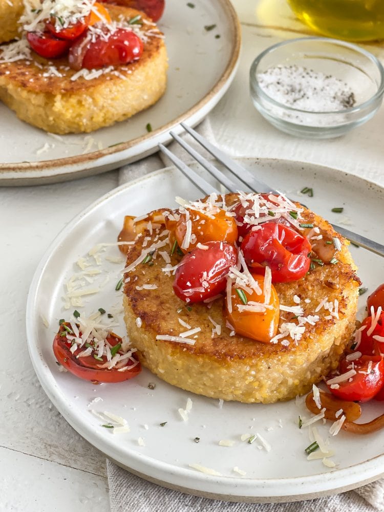 Side view of single polenta cake topped with sautéed cherry tomatoes, parmesan cheese, and chopped rosemary.