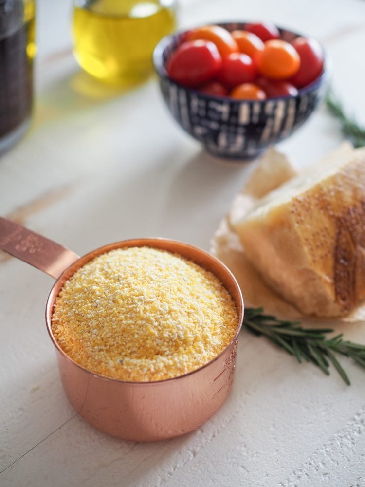 A copper measuring cup filled with course ground yellow cornmeal. A bowl of cherry tomatoes, jar of olive oil, sprigs of rosemary, and chunk of Parmesan cheese in the background.