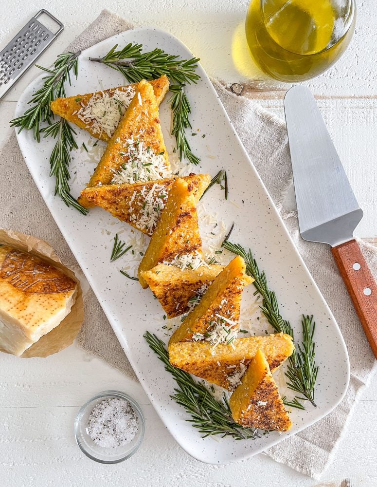 Overhead view of triangle shaped polenta cakes on a rectangular platter, dusted in chopped rosemary and Parmesan cheese. 
