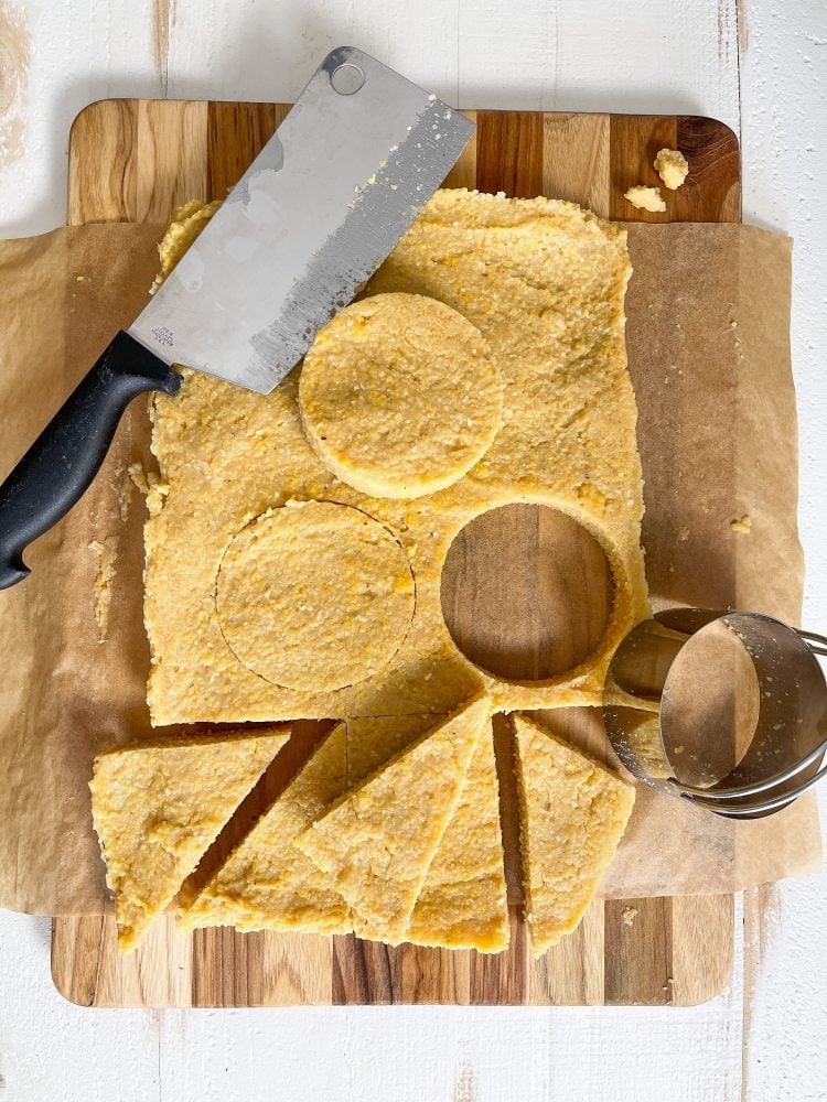 Overhead shot of chilled polenta on a cutting board with different shapes being cut including circles with a biscuit cutter and triangles cut with a sharp knife.