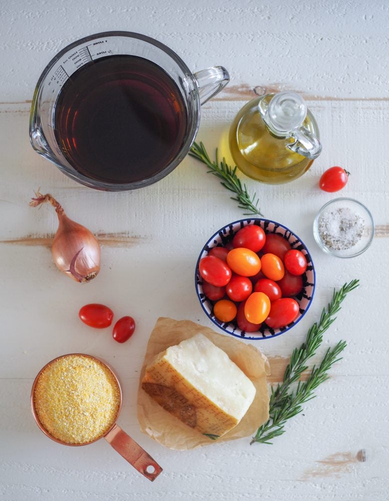 Overhead shot of all the ingredients used in making polenta cakes including vegetable broth, olive oil, rosemary, salt and pepper, shallot, cherry tomatoes, Parmesan cheese, and course ground cornmeal.