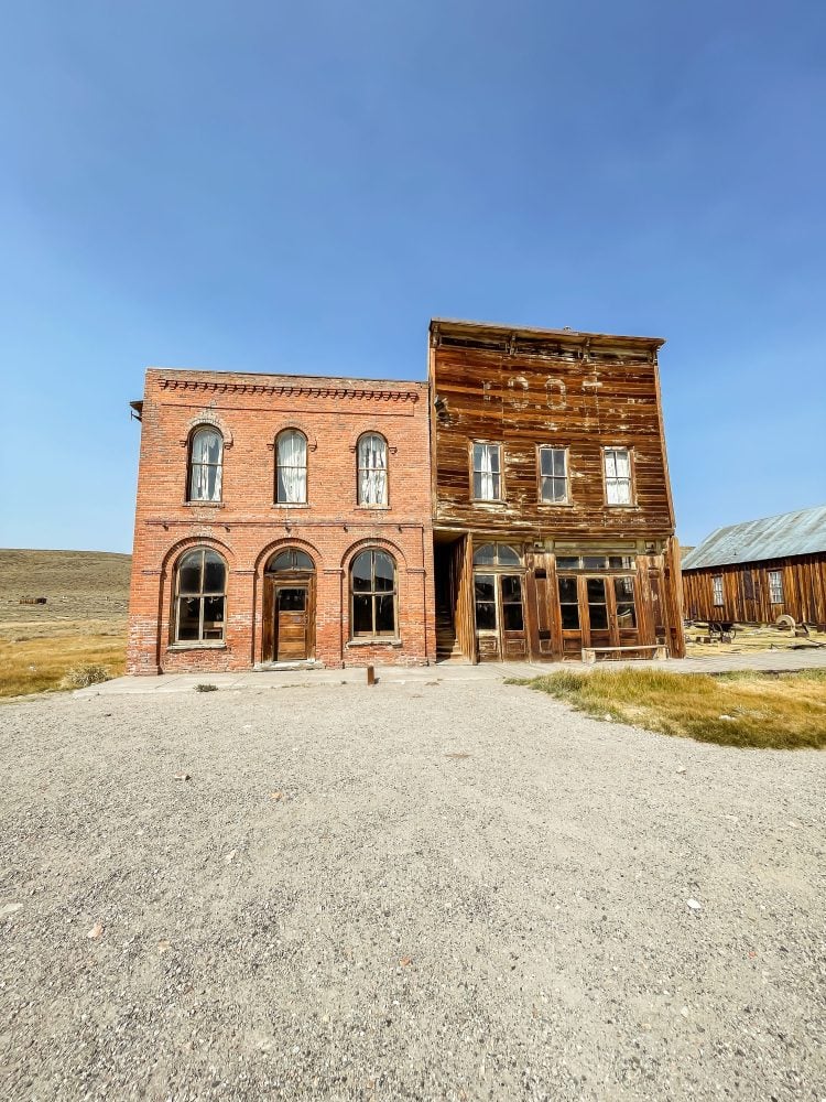 Two old buildings in Bodie ghost town. One on the left is brick with rounded windows. The one on the right is wood and in decay.