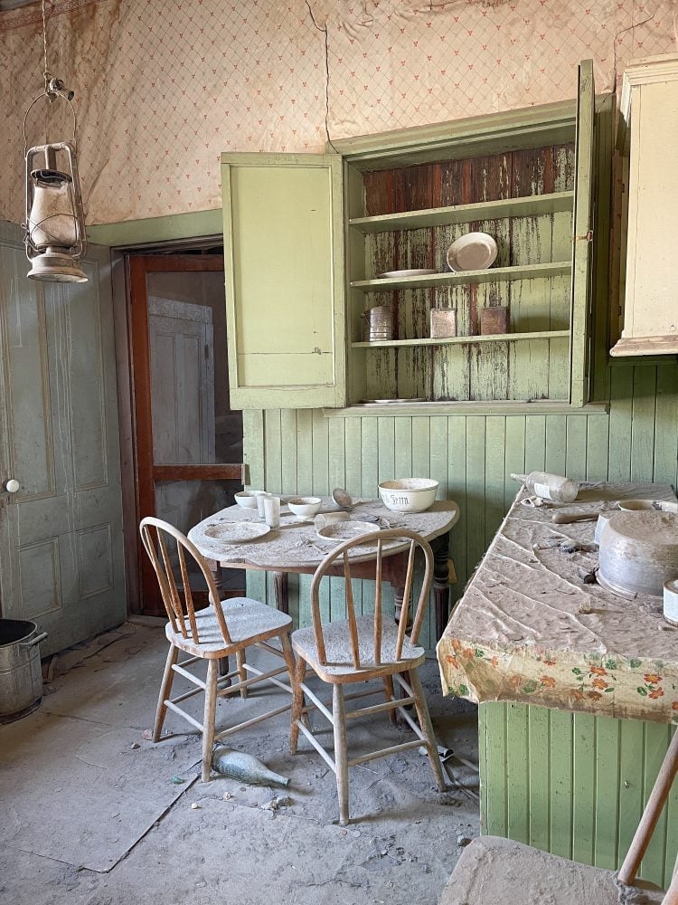 The kitchen inside the Miller House showing a dusty tablecloth on the counter, plates and tins in the pantry, wallpaper and green paint trim, and dishes set on the table. This old house was once a home.