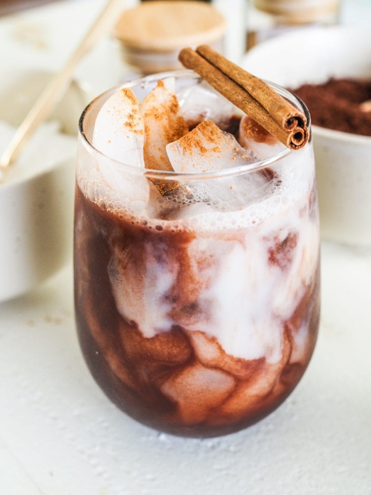 Mexican mocha over ice with milk swirling through it in a glass and topped with a cinnamon stick for garnish.
