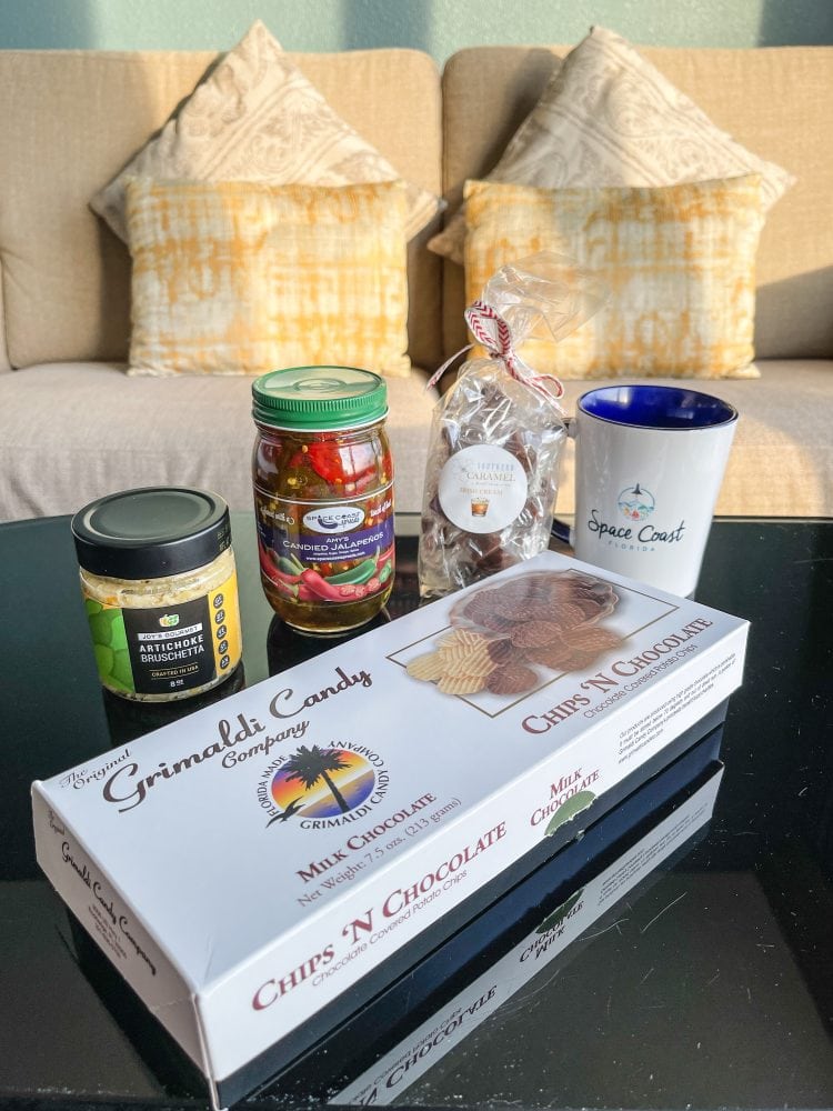 Table with local Space Coast food products including Chips 'N Chocolate, Articoke Dip, Candied Jalapeños, Caramels, and a Space Coast branded mug.