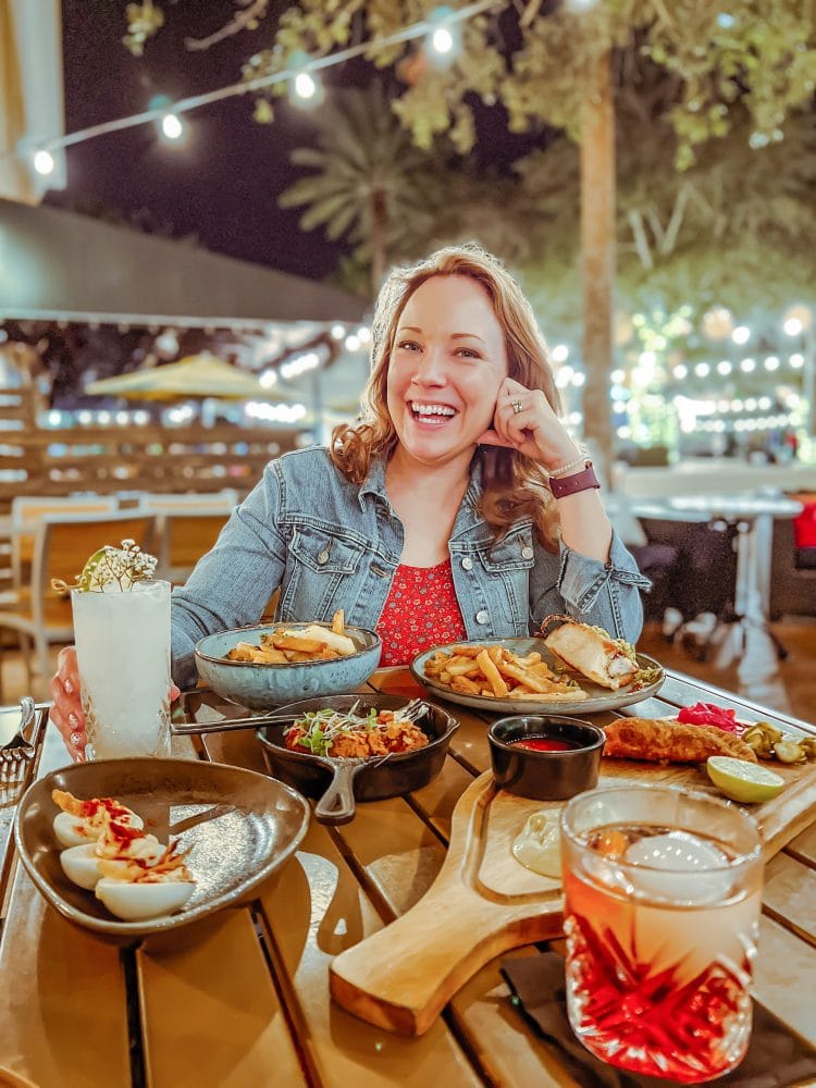 Rachelle sitting in front of table full of appetizers at 28 N Gastro Pub on their outdoor patio with market lights.