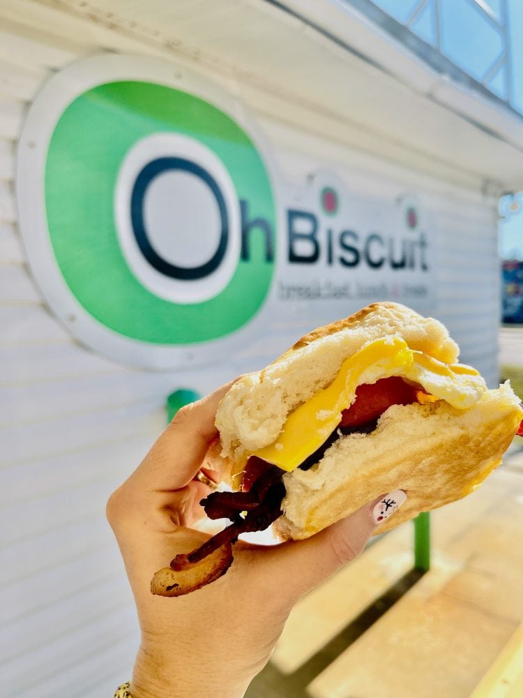 Bacon, egg, cheese and tomato biscuit sandwich held up in front of Oh Biscuit sign.