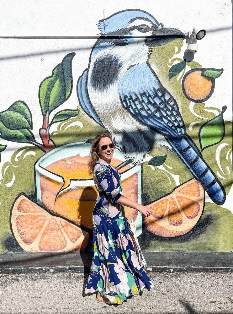 Painted mural on wall of bluejay looking right while standing on a glass of orange juice and orange slices. Rachelle walking by and smiling in front of the mural.