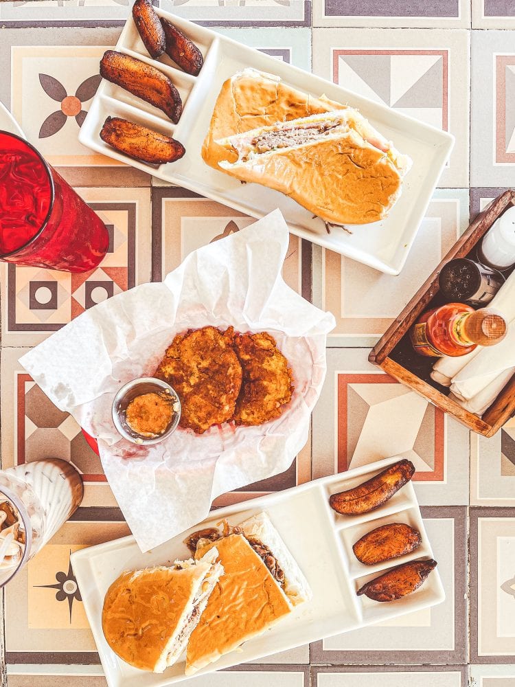 Overhead view of lunch at El Ambia including two pressed sandwiches, fried sweet plantains (maduros) and fried green plantains (tostones) with dipping sauce. 