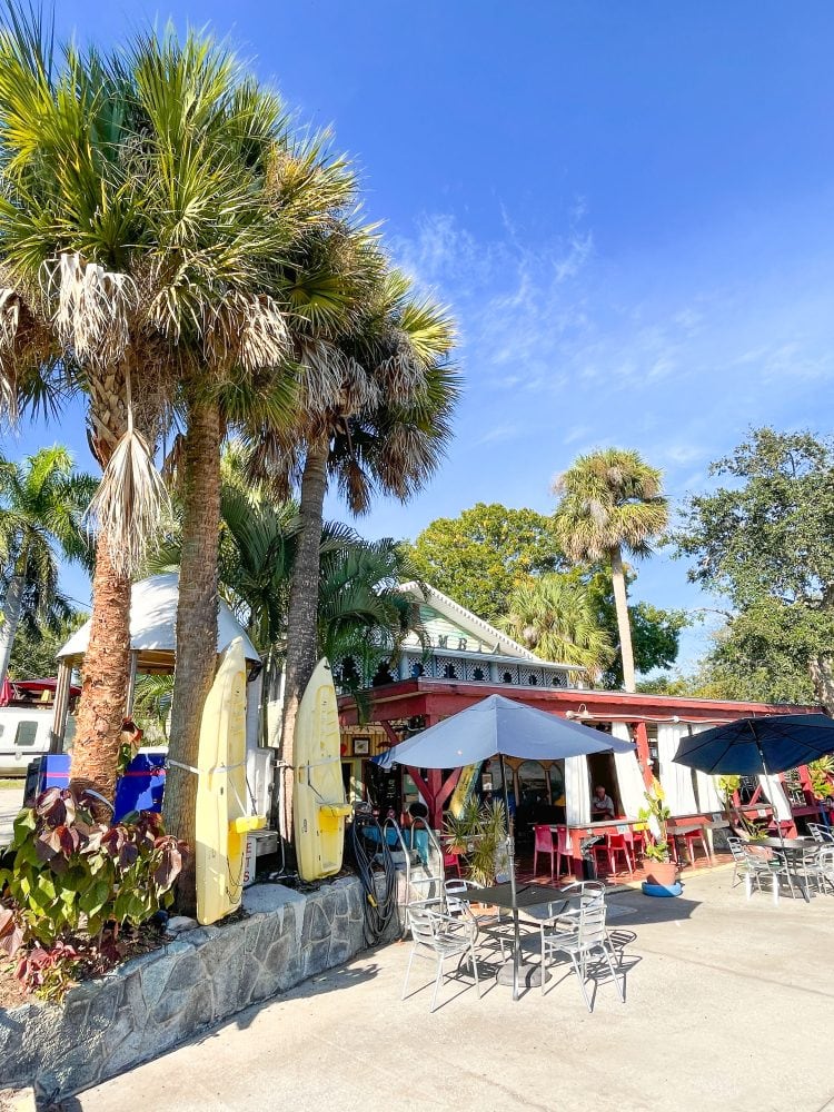 Outside view of El Ambia restaurant with palm trees, decorative kayaks, and tables with blue umbrellas.