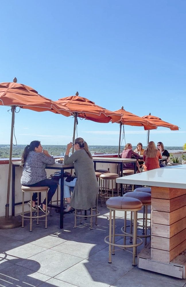 Women sitting at high top tables under orange umbrellas with a rooftop view of Melbourne at the Melby Hotel.