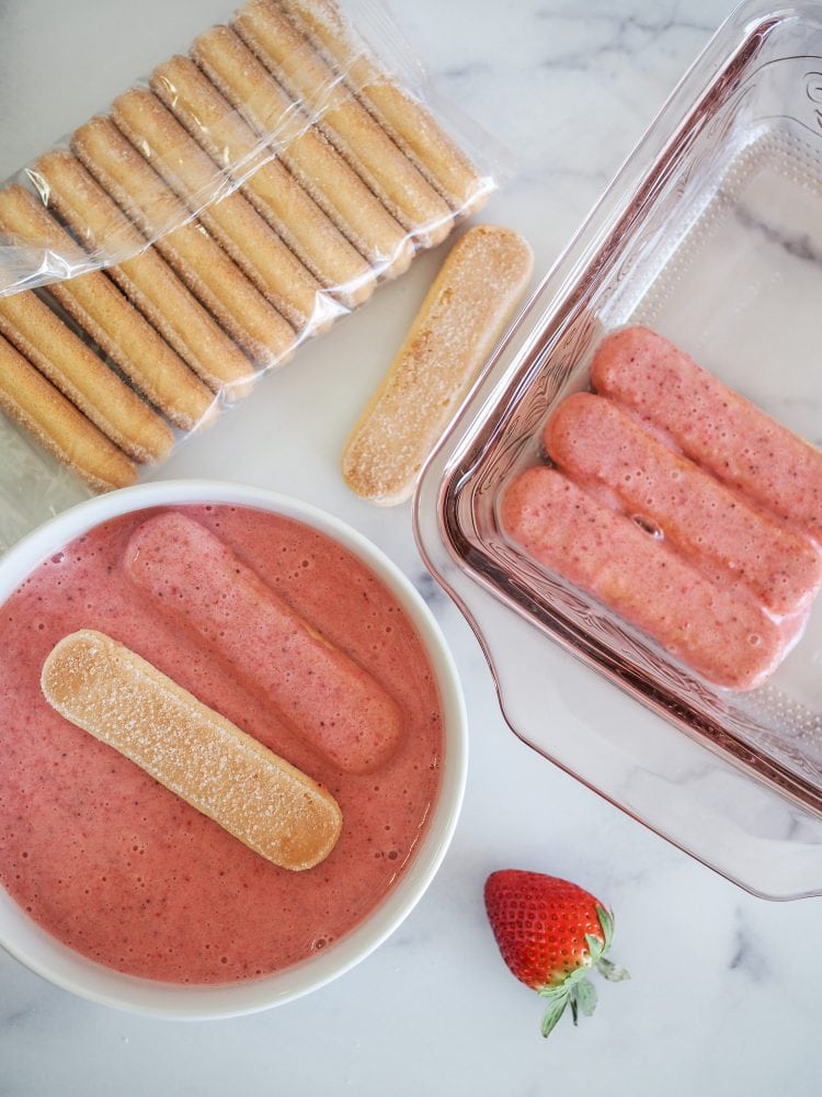 White bowl with two lady fingers soaking in strawberry liqueur puree with package of lady fingers and pink baking pan with first layer of lady fingers starting for strawberry tiramisu.