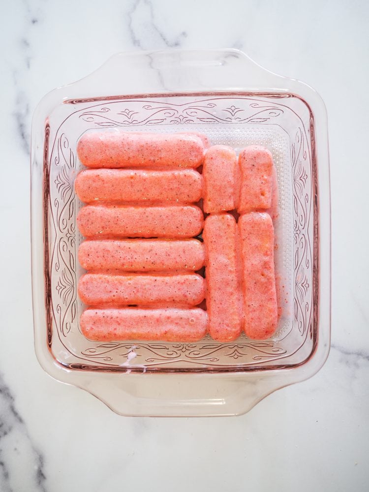 Lady fingers pink with strawberry liqueur puree layered onto the bottom of a clear pink baking pan.