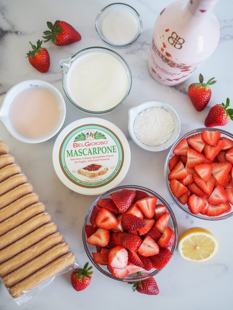 Overhead shot of ingredients to make strawberry tiramisu on a counter including package of lady fingers, tub of mascarpone cheese, Bailey's strawberries and cream, whipping cream, sugar, lemon, and in the foreground a big bowl of halved strawberries.