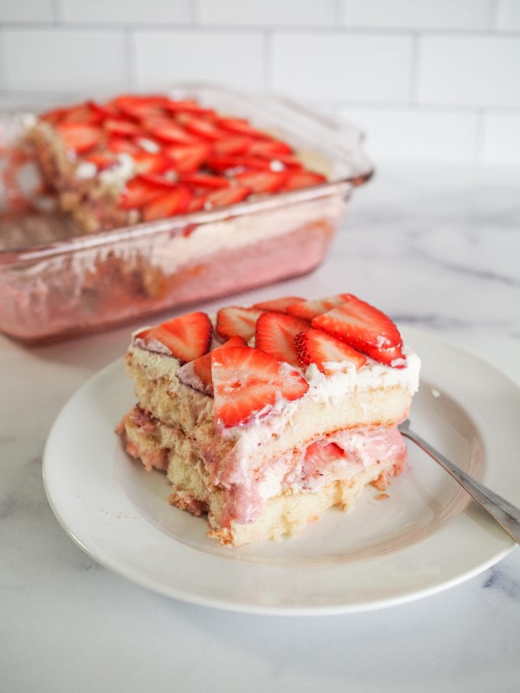 Slice of strawberry tiramisu on a white plate with pink baking dish of tiramisu in background on marble table.