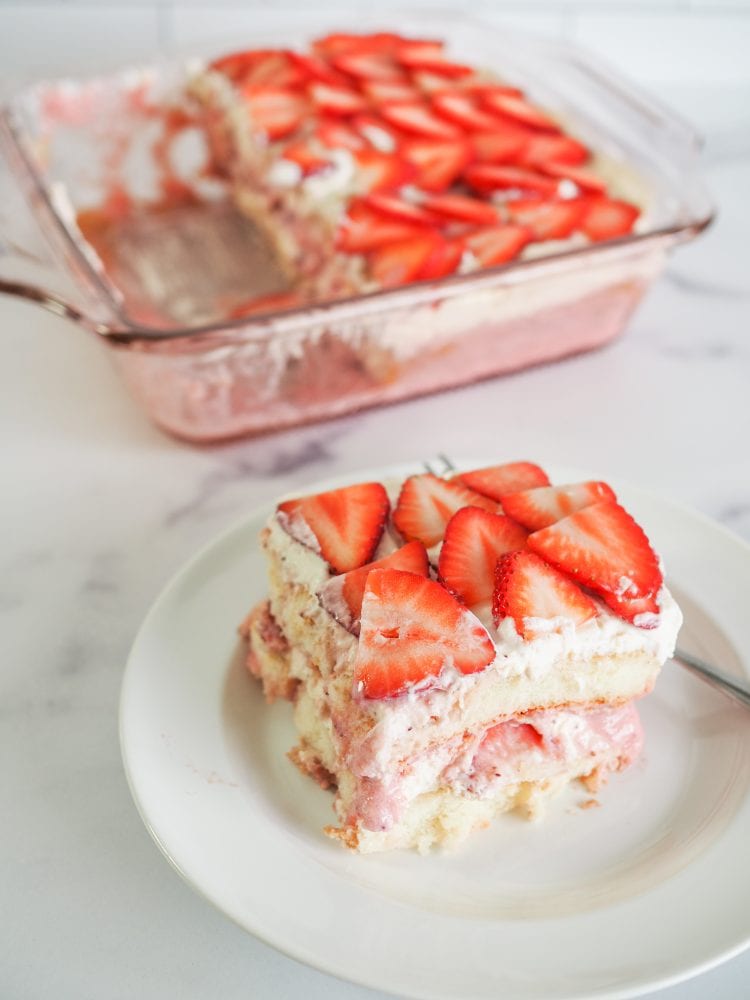 Single slice of strawberry tiramisu on a white plate with pink baking dish with whole strawberry tiramisu in background.