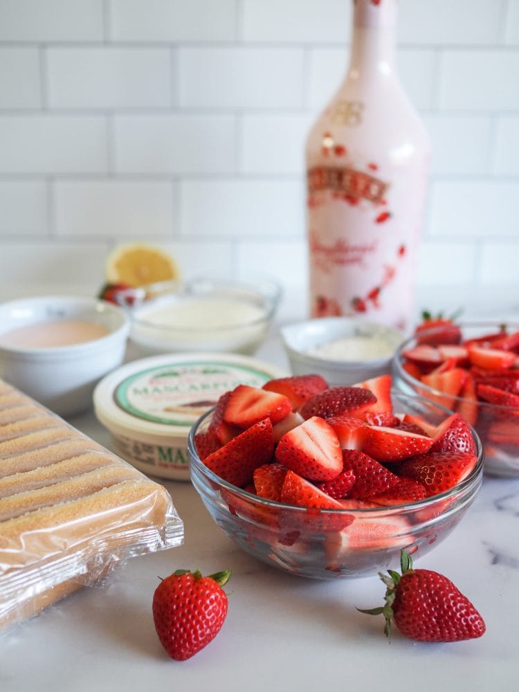 Ingredients to make strawberry tiramisu on a counter including package of lady fingers, tub of mascarpone cheese, Bailey's strawberries and cream, whipping cream, sugar, lemon, and in the foreground a big bowl of halved strawberries.