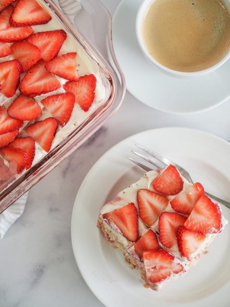 Overhead shot of slice of strawberry tiramisu on a white plate served along side a cup of coffee and whole tiramisu in a pink baking dish on a marble table.