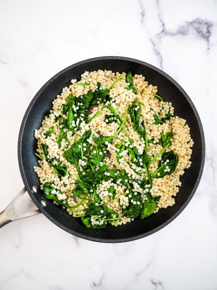 Skillet with cooked couscous, topped with wilted spinach.