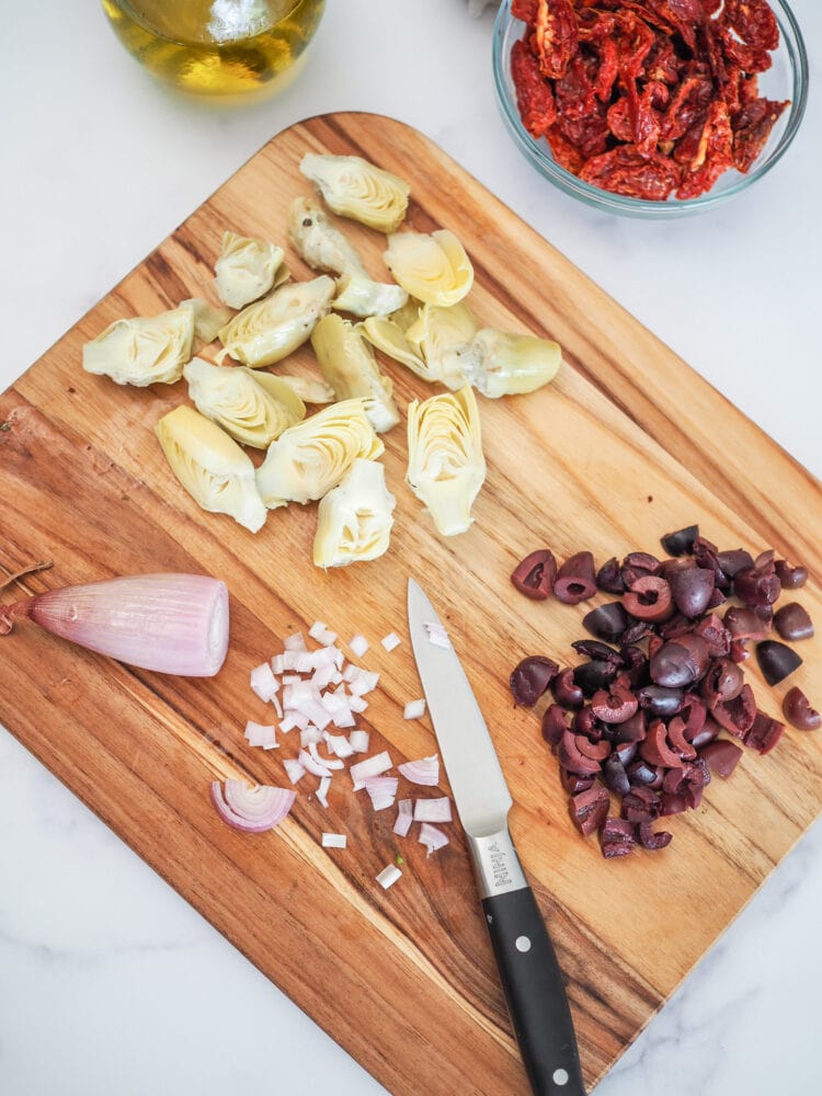 Wood cutting board with olives, baby artchoke hearts, sun dried tomatoes, shallot, and a small knife for chopping.