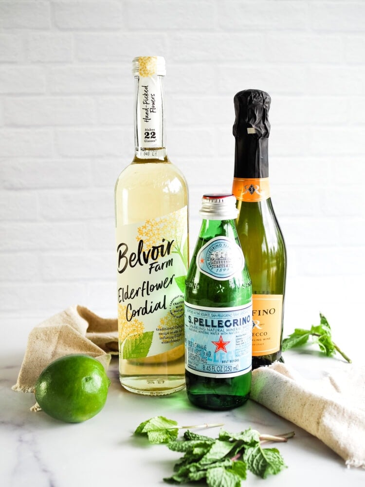 Ingredients to make a Hugo Spritz including a bottle of elderflower liqueur, a bottle of San Pellegrino sparkling mineral water, a bottle of Italian Prosecco, natural linen cocktail napkins, a whole lime, and sprig of mint. 