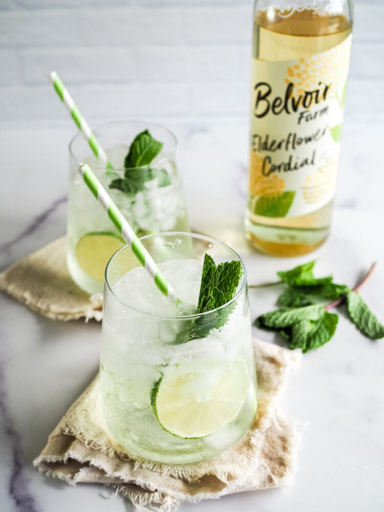 Hugo Spritz cocktail in a flat bottomed stemless wine glass with a slice of lime, sprig of fresh mint, and green and white striped straw. The photo is angled looking into the glass and there's a bottle of elderflower syrup in the background against a white brick backdrop.