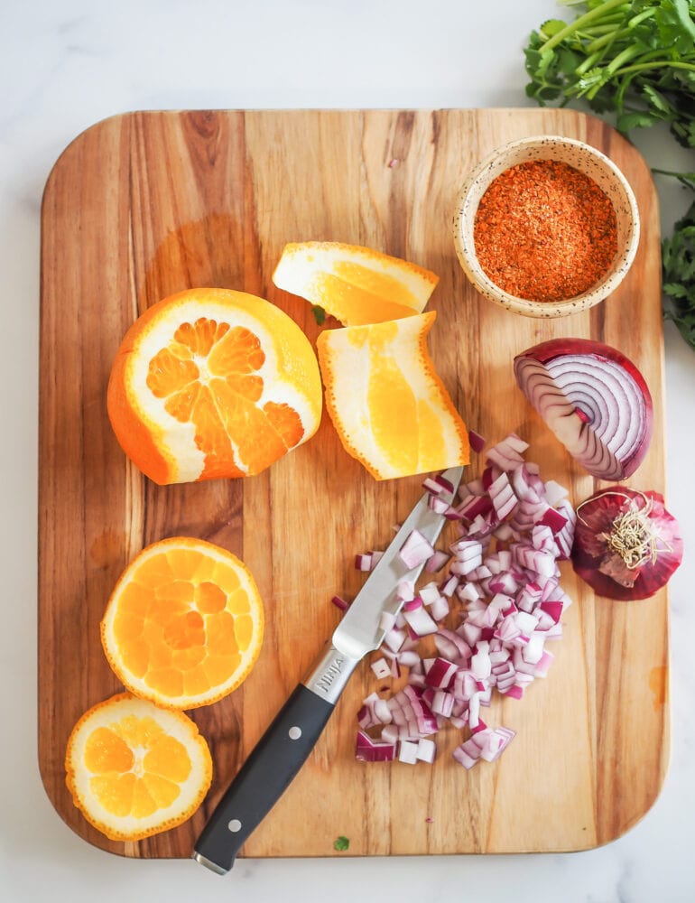 Cutting board with orange (peels sliced away), diced red onion, and tiny prep bowl filled with chili lime seasoning.