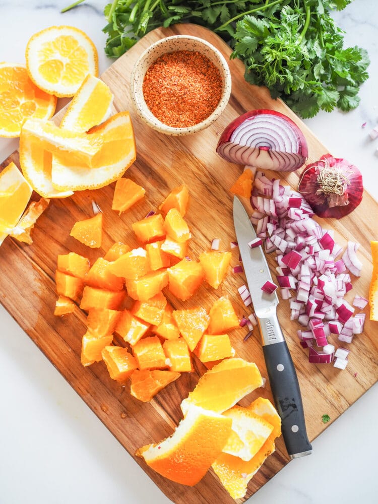 Cutting board with sliced orange, diced red onion, and tiny prep bowl filled with chili lime seasoning.