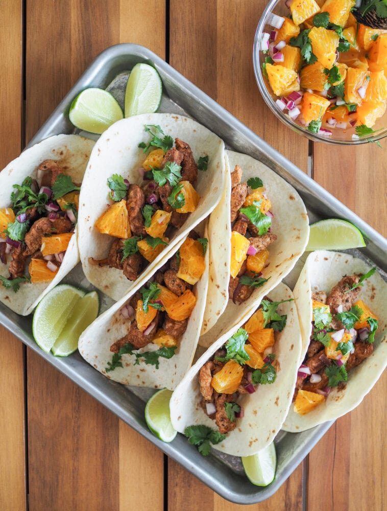 Overhead shot of six pork tacos on a tray, garnished with citrus salsa and lime.