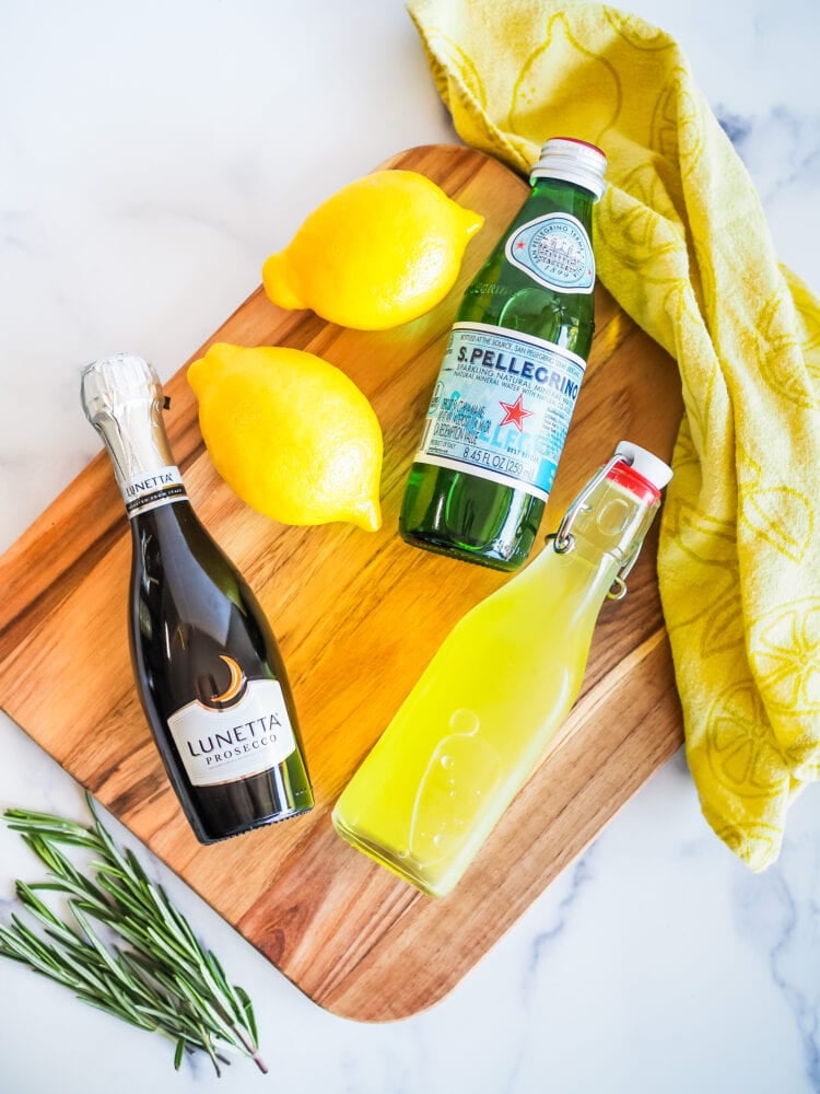 Ingredients for a limoncello spritz: bottle of Prosecoo, bottle of sparkling water, bottle of limoncello, rosemary, and lemons.