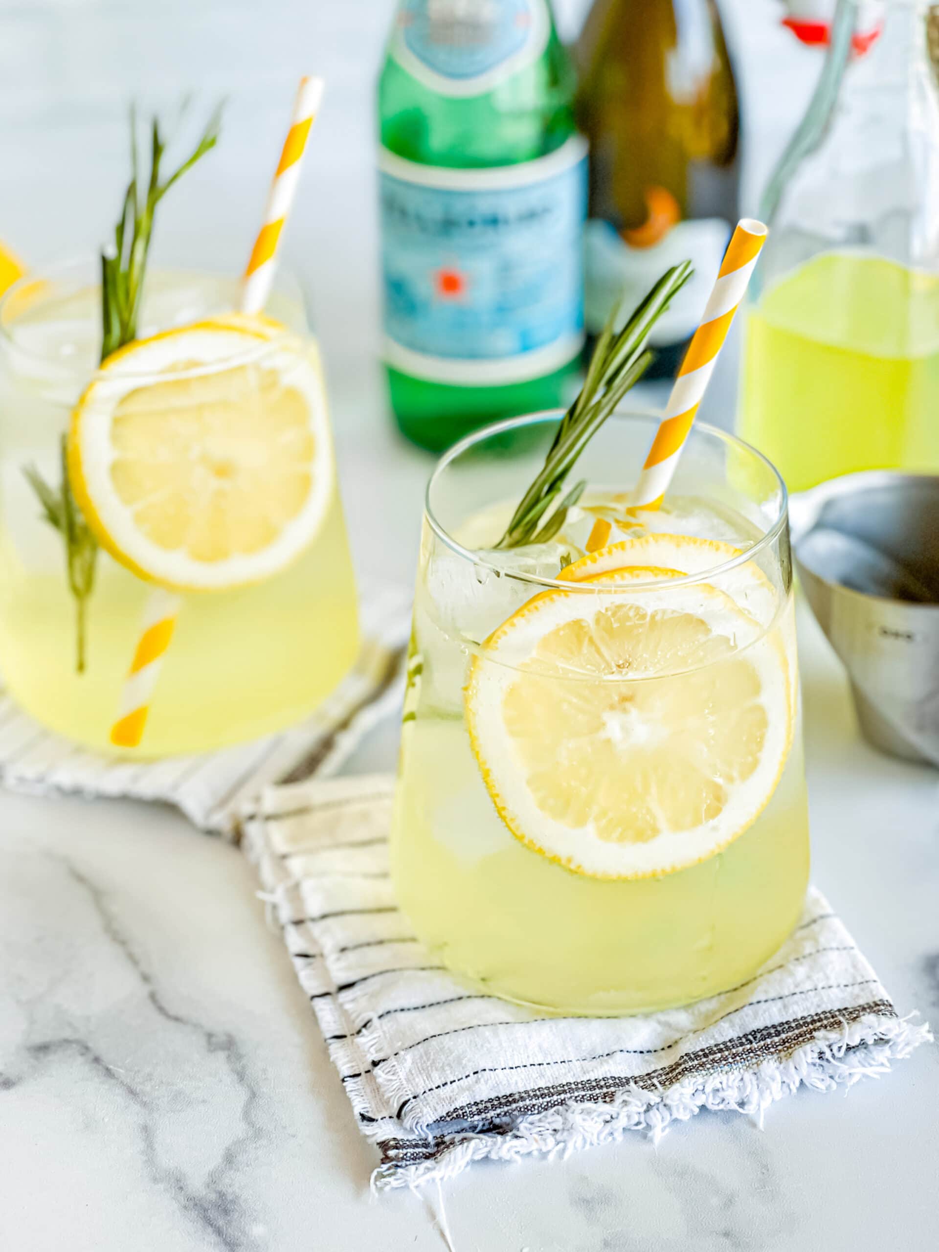 Two glasses of limoncello spritz, garnished with lemon, rosemary, and yellow striped straw. Sparkling water, Processo, and limoncello in the background.