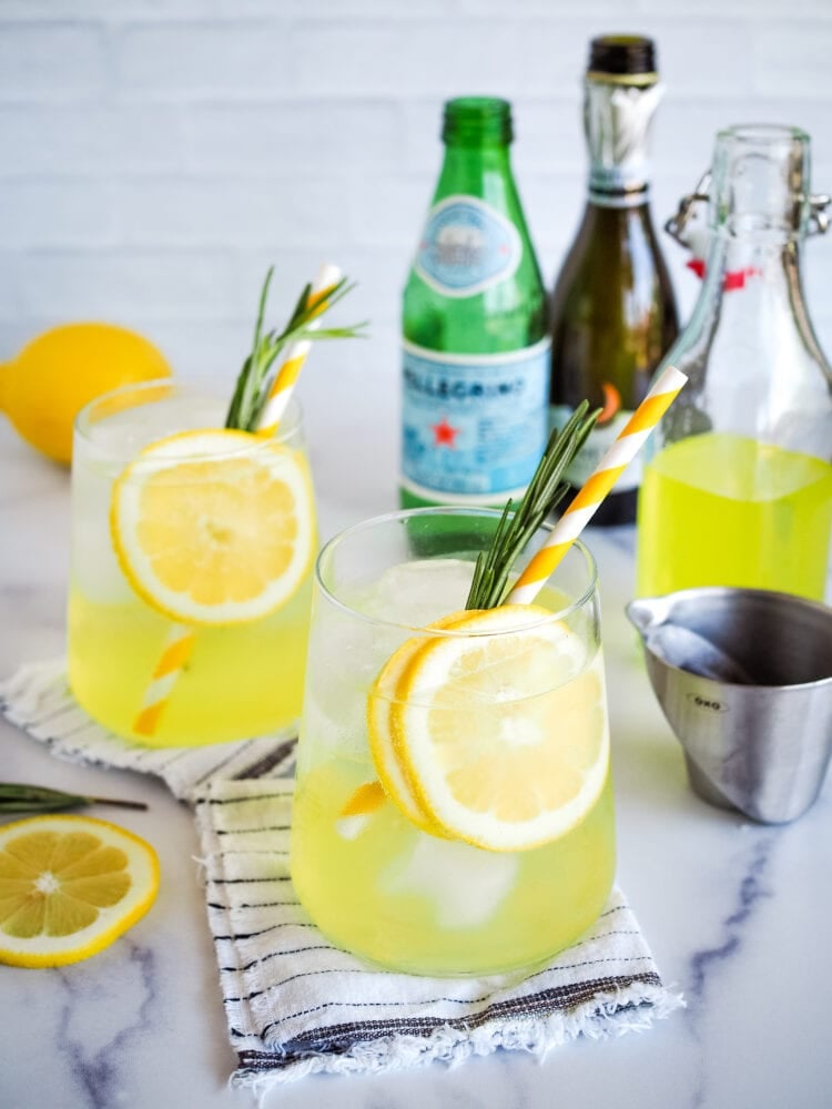 Two glasses of limoncello spritz, garnished with lemon, rosemary, and yellow striped straw. Sparkling water, Processo, and limoncello in the background.