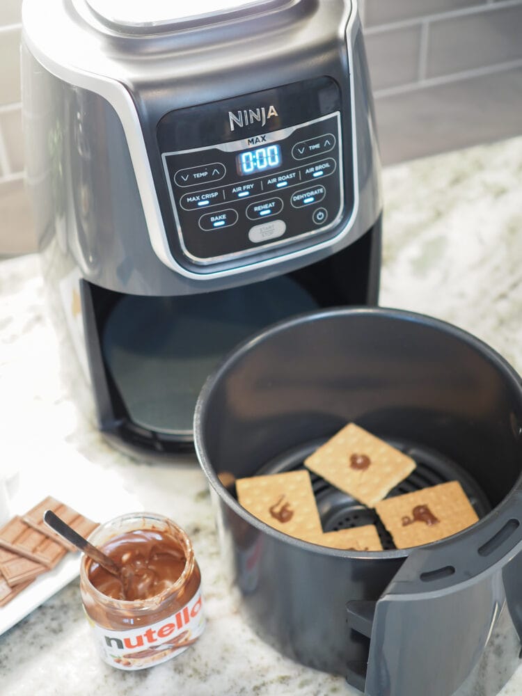 Ninja XL air fryer with four graham crackers inside and a dab of Nutella on each. A jar of Nutella and a plate of Hershey's chocolate is off to the side.