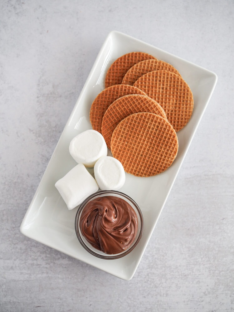 Six stroopwaffels, three marshmallows, and a small dish of Nutella on a serving plate.