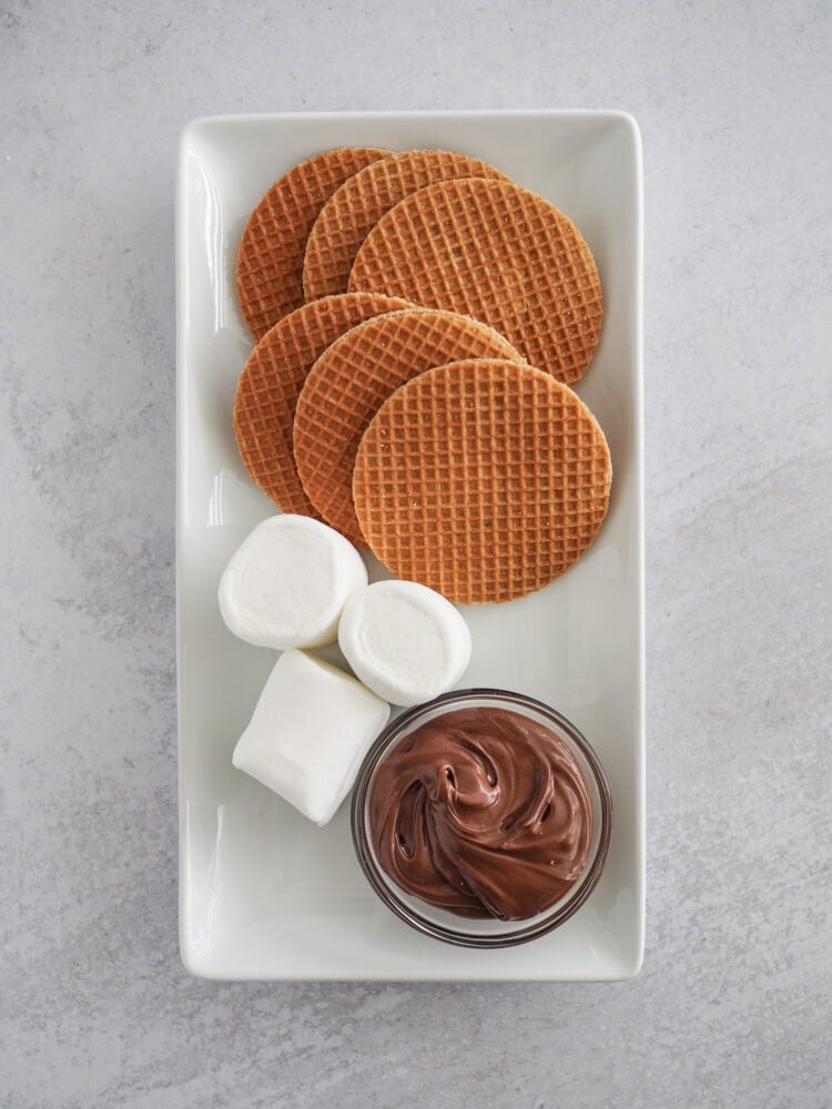 Six stroopwaffesl, three marshmallows, and a small bowl of Nutella on a white serving plate.